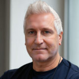 A person with short, light-colored hair and a relaxed expression looks directly at the camera, wearing a dark shirt, against a neutral background.