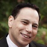 Smiling person in a suit with a blurred leafy background.