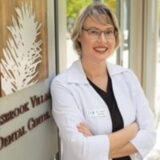 A friendly dentist with glasses in a white coat stands smiling outside a dental clinic.