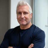 A confident man with short, light-colored hair stands with his arms crossed, wearing a dark sweater. He is smiling gently, conveying warmth and approachability. The background includes soft natural light and a minimalist aesthetic.