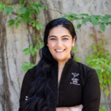 A woman with long black hair stands smiling in front of a green backdrop. She wears a black jacket that features a logo, suggesting she is a dental professional. Her friendly demeanor conveys approachability and confidence.