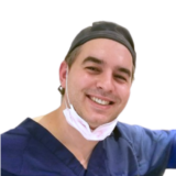 A smiling dentist wearing a blue scrub top and cap, with a surgical mask under his chin, stands against a plain white background.