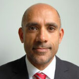 A person wearing a suit and tie is shown with a neutral expression against a plain background.