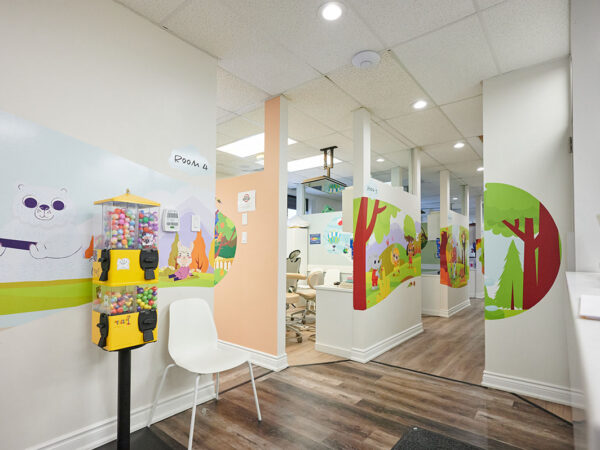 A children's dental office with colorful wall designs, featuring playful forest themes. There's a small candy dispenser and a white chair near the entrance, leading to treatment areas. The atmosphere is bright and inviting for kids.