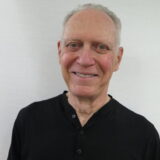A smiling older man wearing a black shirt stands against a plain white background.