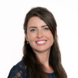 A woman with long dark hair smiles warmly at the camera, wearing a dark lace top, against a plain white background.