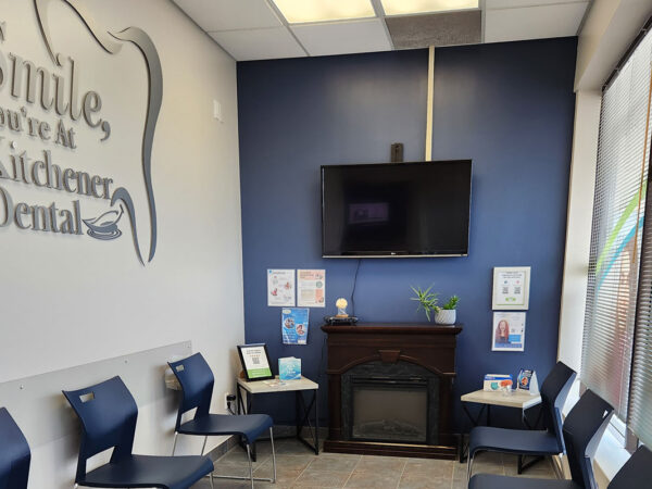 Dental clinic waiting area with blue chairs, a wall-mounted TV, and friendly decor. There's a sign saying "Smile, You're at Kitchener Dental" and a few informational posters. The room is well-lit and welcoming.