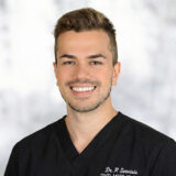 A man with short brown hair and a beard wearing a black V-neck shirt smiles against a blurred background.
