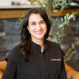 A smiling person wearing a black dentist coat with "Dr. Maria Syed" embroidered, sitting in a modern office with plants and a stone wall in the background.