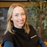 A smiling woman with blonde hair wearing a dark jacket sits in a warmly lit room with plants in the background.