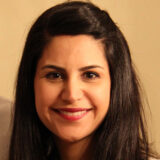 A woman with long dark hair smiles warmly at the camera. She has an approachable demeanor, exuding confidence and friendliness.