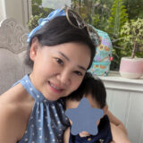 A smiling woman in a blue polka dot dress poses for a photo with a small child. The child’s face is obscured with a star shape. In the background, there is a playful backpack and decorative plants. The friendly 123Dentist Smiley adds a cheerful touch to the scene.