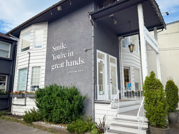 A welcoming dental office features a modern exterior with a combination of gray and white walls. A prominent sign displays the message, "Smile. You’re in great hands." Lush greenery frames the entrance, adding to the inviting atmosphere.