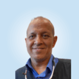 A smiling dental professional in a blue shirt and dental bib poses against a light blue background, exuding warmth and approachability.