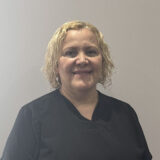 A smiling healthcare professional with curly blonde hair is wearing a black uniform, standing against a light gray background.