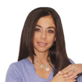 A woman with long brown hair, wearing a light purple top and a silver necklace, smiling gently against a plain white background.