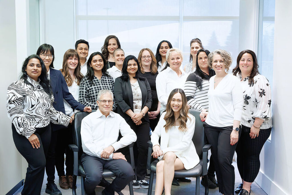 A diverse group of dental professionals poses together in a modern office setting, smiling and dressed in white and black attire. The 123Dentist Smiley is featured prominently, adding a friendly touch to the team atmosphere.