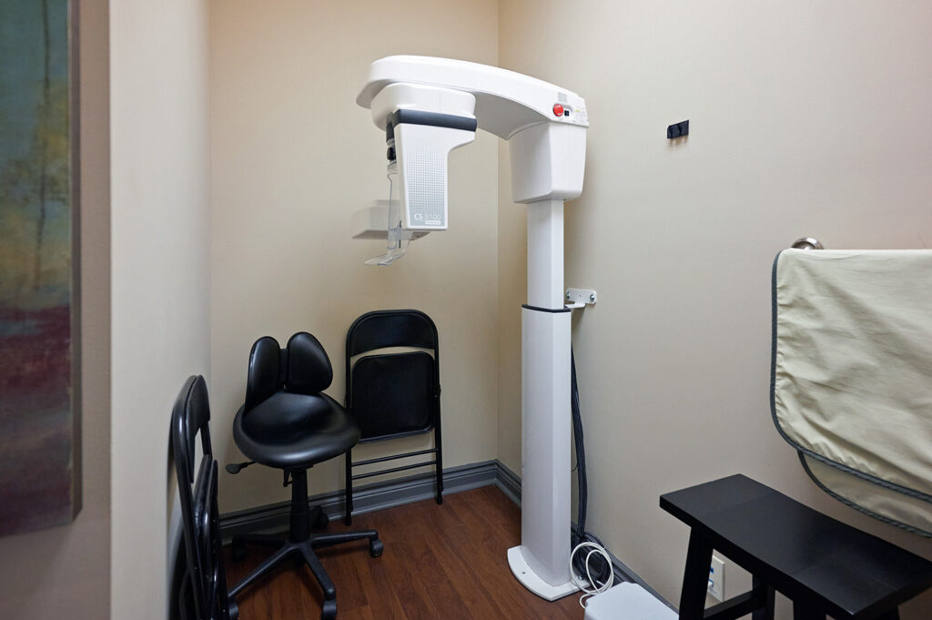 A dental examination room featuring a digital X-ray machine, two chairs, and a privacy screen. The space is clean and well-lit, ready for patient use.