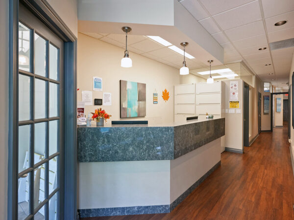 A dental office interior featuring a reception desk with a stone countertop, brightly lit by ceiling fixtures. The walls are painted in light colors, decorated with artwork, and a warm wood floor adds to the inviting atmosphere. The 123Dentist Smiley adds a friendly touch.