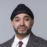 A person with a beard wearing a black turban, a plaid suit jacket, a white shirt, and a patterned tie poses against a gray background.