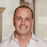 A smiling man with short hair is wearing a white shirt and standing in a bright, modern environment. He appears friendly and approachable, embodying a welcoming demeanor.