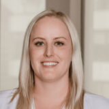 A smiling woman with blonde hair, wearing a white top, stands in a well-lit indoor setting. The background features soft, blurred elements, suggesting a professional environment.