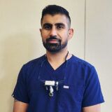 A male dentist wears a navy blue scrubs top and dental loupes around his neck, smiling confidently at the camera.