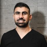 A person with short dark hair and a beard, wearing a black V-neck shirt, smiles against a textured gray wall.
