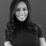 Smiling woman with long hair wearing a dark top, posed against a plain background.