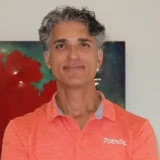 A person with gray hair wearing an orange shirt with the text "7Dental," stands in front of a colorful background.