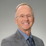 A smiling man wearing glasses and a gray plaid suit stands against a neutral background. He has short, light-colored hair and exudes a friendly, approachable demeanor.