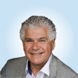 A smiling man with curly gray hair wearing a gray checkered blazer and white shirt, set against a light blue background.