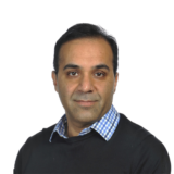 A person with dark hair wearing a checkered shirt and a black sweater, set against a plain white background, looks directly at the camera with a neutral expression.