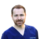 A person wearing blue medical scrubs, with a friendly expression and short brown hair, against a white background.