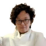 A person with short curly hair and glasses, wearing a white jacket, is looking directly into the camera against a plain white background.