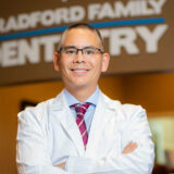 A smiling dentist stands confidently with arms crossed, wearing a white coat and a tie. The background features a sign that reads "Bradford Family Dentistry," conveying a warm and professional dental environment.