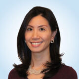 A smiling woman with long, dark hair and earrings poses against a light blue background. She wears a dark top and a decorative necklace, exuding a warm and friendly demeanor.
