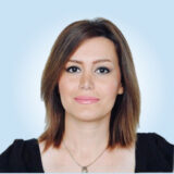 A person with shoulder-length brown hair and a neutral expression, wearing a black top, is posed against a light blue background.