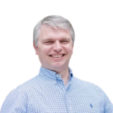 A person with gray hair, smiling broadly, wearing a blue checkered shirt against a plain white background.