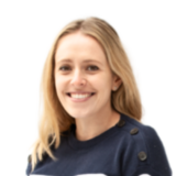 A smiling woman with long blonde hair wearing a navy sweater with buttons on the shoulders, set against a plain white background.