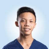 A person in a blue scrub top is smiling at the camera against a light blue background.