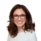 A person with shoulder-length brown hair and glasses is smiling warmly, wearing a light-colored top against a plain white background.