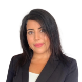 A person with long dark hair wearing a black blazer over a white top, smiling slightly, against a white background.
