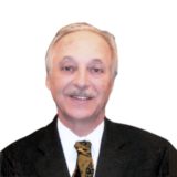 A man with light hair and a mustache is wearing a dark suit and patterned tie, smiling against a plain white background.