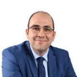 A man wearing glasses and a blue suit smiles warmly at the camera against a plain white background.
