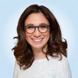 A smiling person with brown hair and glasses, wearing a white top, against a light blue background.