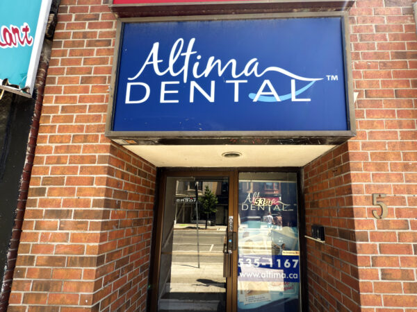 A dental office entrance featuring a blue sign that reads "Altima DENTAL" above the door. The exterior is made of red bricks, and the door is glass, allowing a view of the interior space.