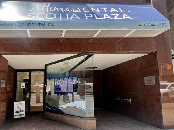 The image shows the entrance of a dental clinic named "Altima Dental, Scotia Plaza." The storefront features a modern design with a prominent sign displaying the clinic's name and contact information. Bright lighting enhances the welcoming atmosphere.