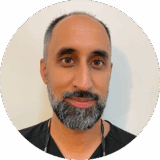 A smiling individual with a beard, wearing a dark shirt, poses against a light background. The image is circular in shape.