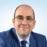 A professionally dressed man in a blue suit and glasses smiles warmly against a light blue background, conveying a sense of friendliness and approachability.